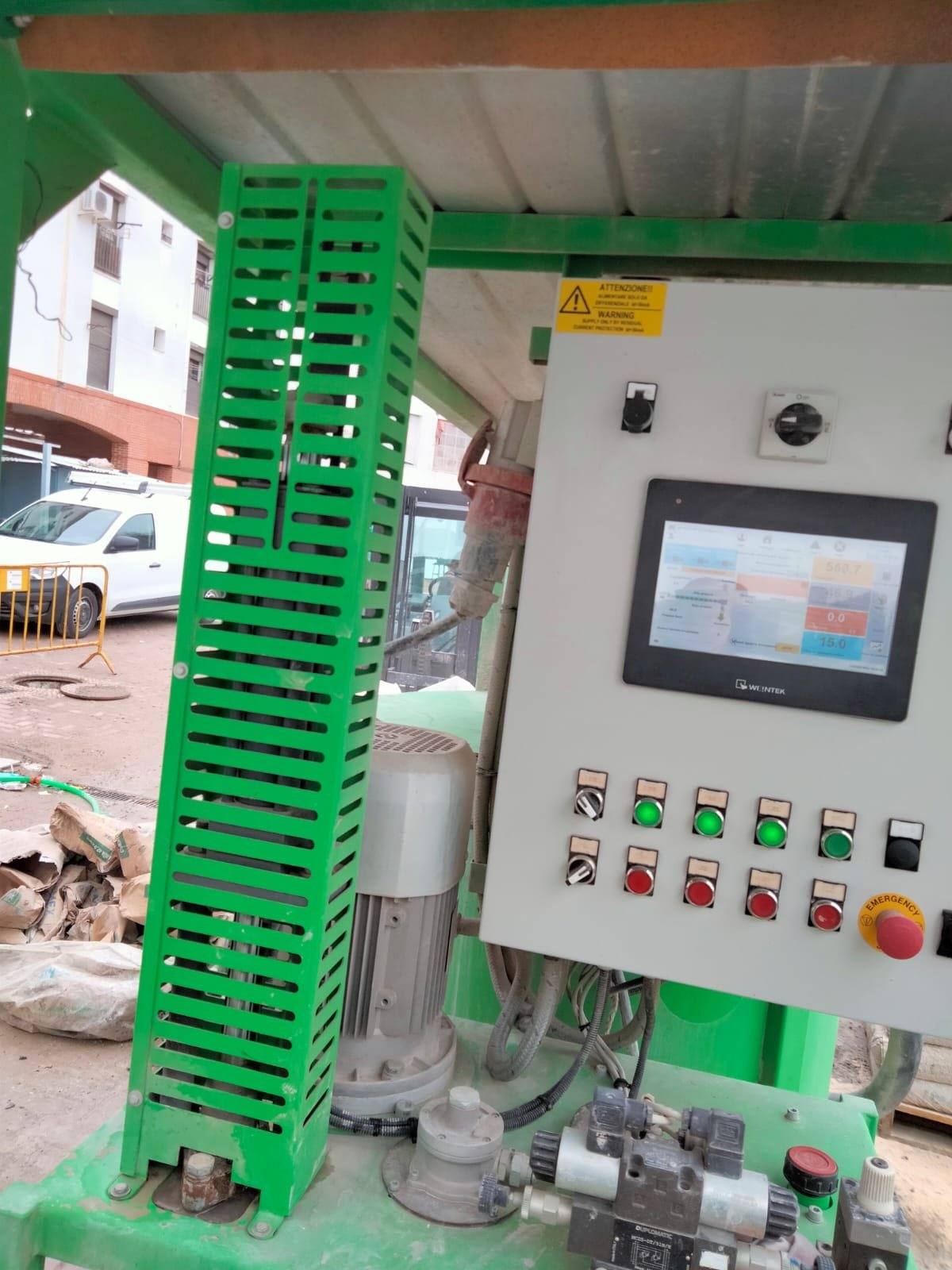 Green grouting plant with its control panel: touch screen, green and red push buttons and emergency stop, beside bags of cement.