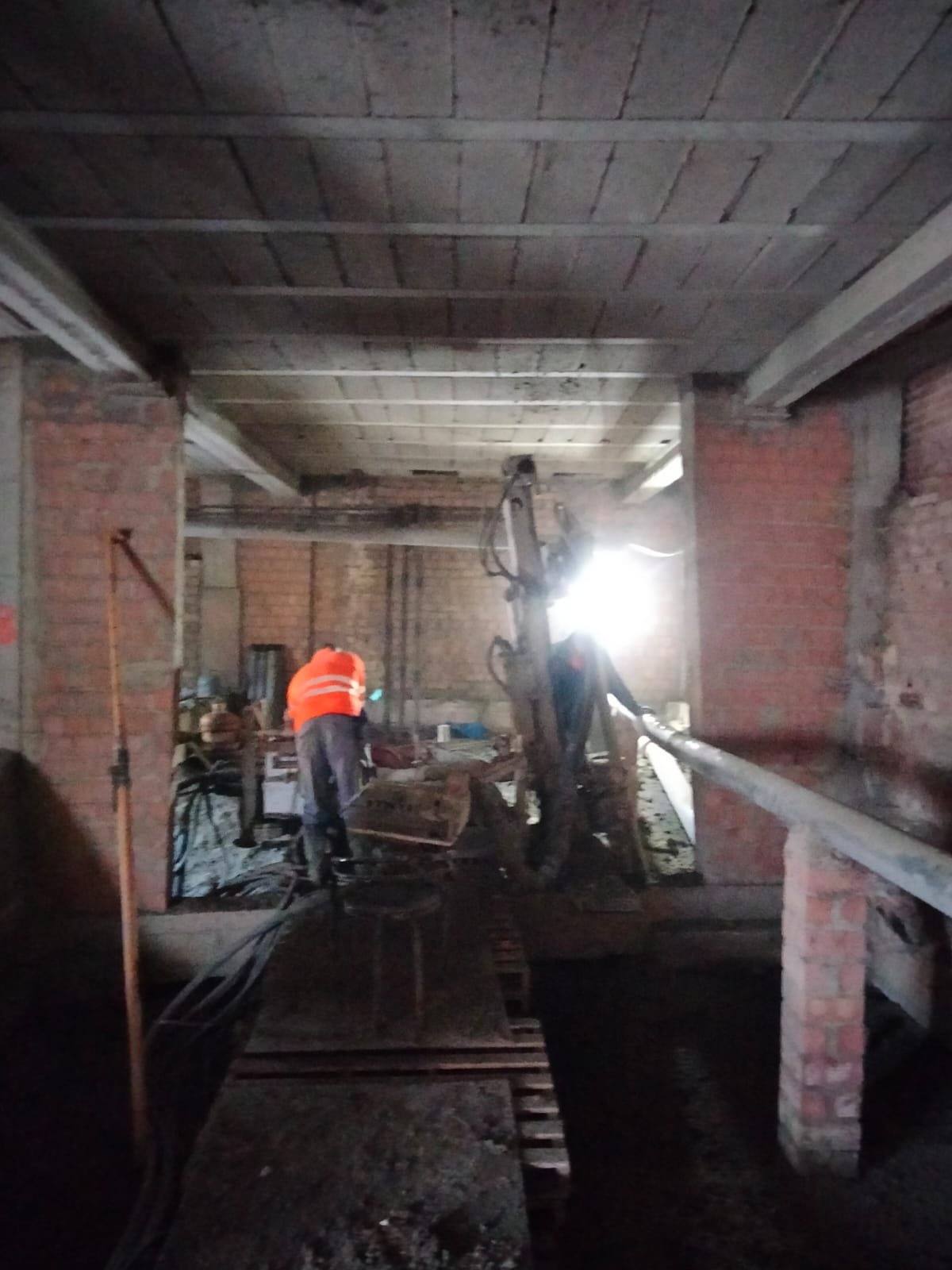 Operative in an orange vest beside a drilling rig lit by a floodlight, inside a stripped-out ground floor, between brick columns and beneath the floor slab.