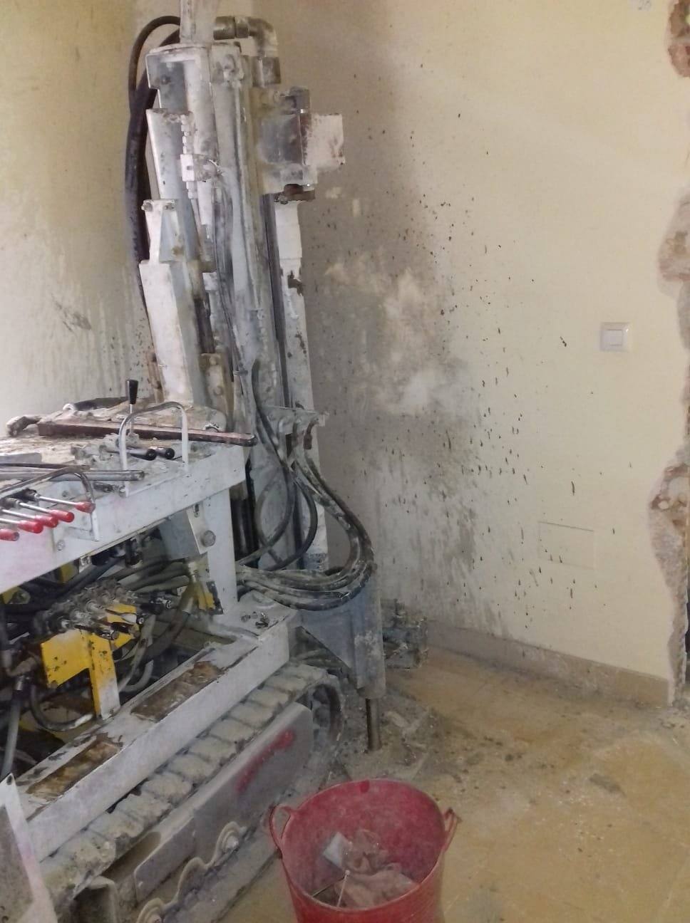 Perforadora de mástil vertical junto a un muro interior, con el pavimento levantado y la pared protegida.